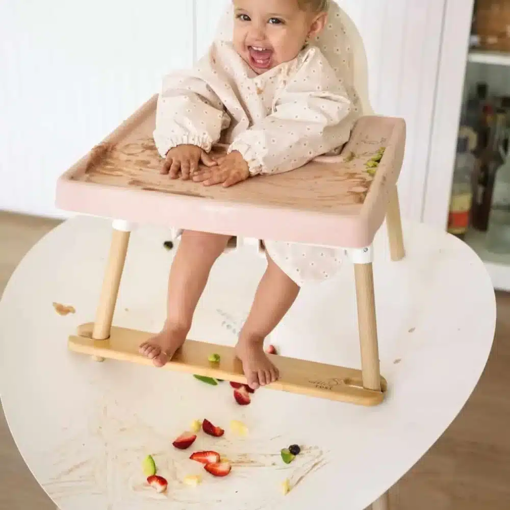 A happy child sits in a highchair with a Nibble and Rest Messi Highchair Food Catcher attached around the legs to catch dropped food.