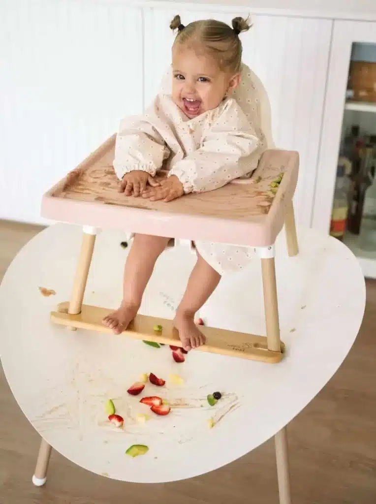 A happy child sits in a highchair with a Nibble and Rest Messi Highchair Food Catcher attached around the legs to catch dropped food.