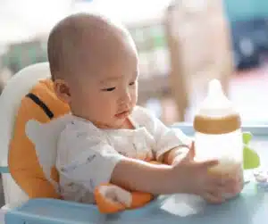 Asian Baby in a highchair holding a bottle of milk.